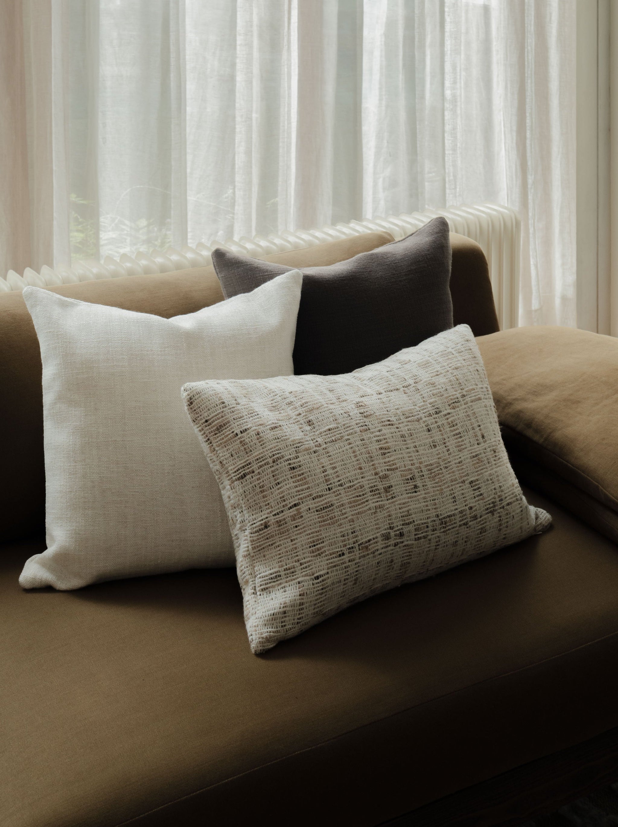 Raw Tones – The Asymmetric Trio, featuring three textured cushions in warm neutral tones, styled on a dark sofa