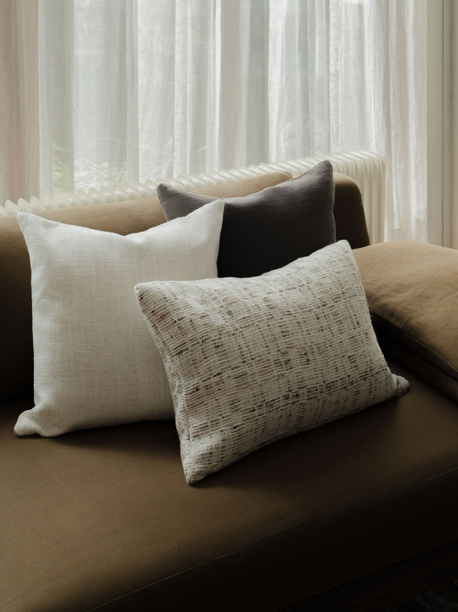 Raw Tones – The Asymmetric Trio, featuring three textured cushions in warm neutral tones, styled on a dark sofa